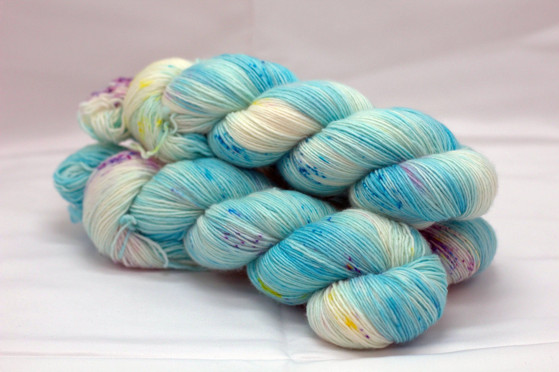 Side view of three twisted skeins variegated turquoise and white yarn with yellow and purple speckles on white background.
